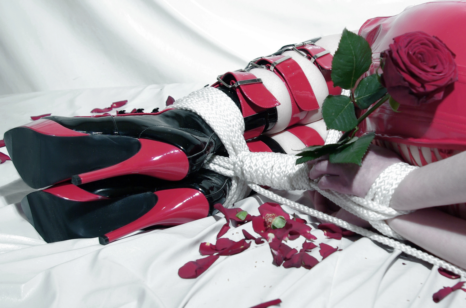 Woman wearing a red dress and black and red heels bound with rope in a shibari tie holding a rose and surrounded by rose petals